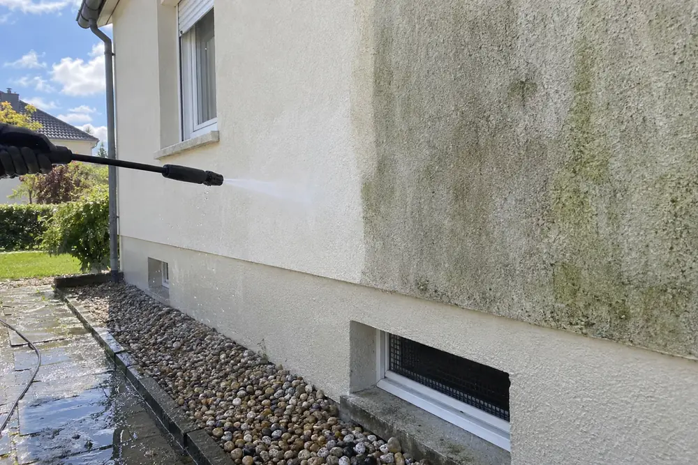 Façade en enduit clair en cours de nettoyage à la lance haute pression : une bande lavée, nette, à côté de l’enduit encore verdi